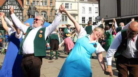 The Shrewsbury Morris Dancers performing in the town. The women are wearing colourful pinafore dresses over white tops and the men are wearing white shifts with green Morris dancing jackets. They are dancing in a circle, facing the outside with their backs to the centre. The town, including a black and white building, is in the background.