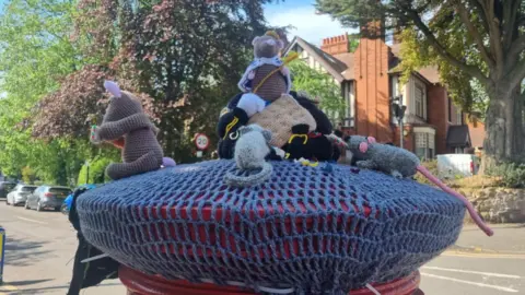 Moseley Knit and Natter Group A knitted rat, with a crown and a sceptre designed to look like cheese, sits on top of a mound of knitted bin bags on top of a postbox. Other knitted rats can be seen sat around it.