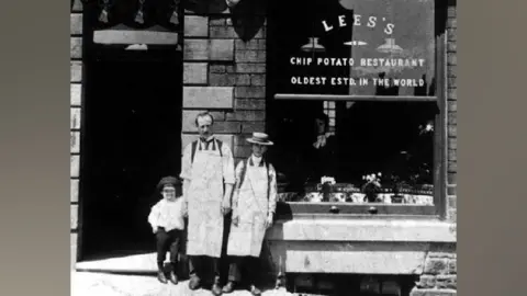 Tameside Image Archive Black and white photo showing a child, man and boy standing in front of Lee's fish and chip shop