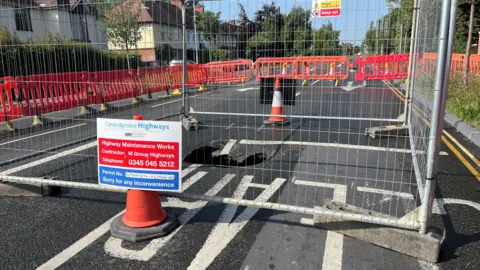 Tom Jackson/BBC A sinkhole is in the middle of a road which has been closed. Around the sinkhole is a metal fence and cones with signs. Surrounding that are red barriers which have closed the road.
