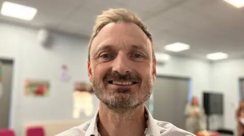 Shariqua Ahmed/BBC Matt Oliver se s úsměvem dívá přímo do kamery. Má na sobě košili s bílým vzorem a má krátké hnědé vlasy a plnovous. 