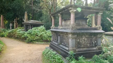 Two old tombs in Highgate Cemetery. They have weathered with age and have columns so the graves stands high off the ground. They are surrounded by greenery and a path. 