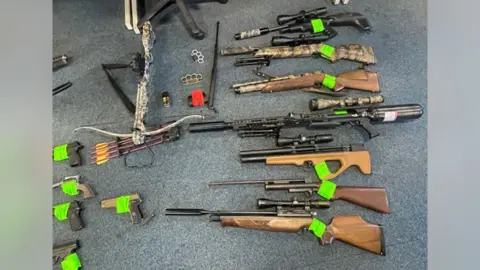 Many weapons laid out on a grey carpet floor. Seven air rifles are in a vertical line to the left of th frame. A crossbow and four arrows are to the right. Just out of frame are what appear to be five handguns. Two knuckledusters are in the middle of the frame. A baton is also among the pile.