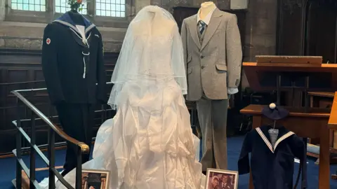 Carol Atkinson A navy uniform, a grey three-piece suit and a white wedding dress with an extravagant veil are worn on mannequins. The items are displayed in a church pew as part of an exhibition.