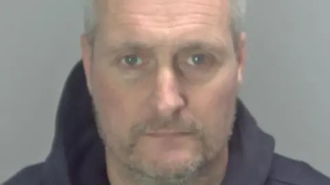 Oliver Arnold has short grey receding hair and a stubbly beard and moustache. He appears on this police custody photo wearing a blue hooded top. The background is grey.