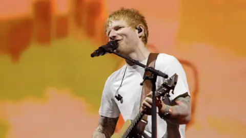 Ed Sheeran in a white shirt playing the guitar with a hazy orange background behind him