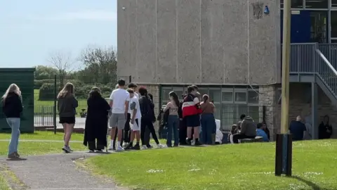 Queue of students heading in to get the antibiotics at a school in Weymouth. It is a sunny day.