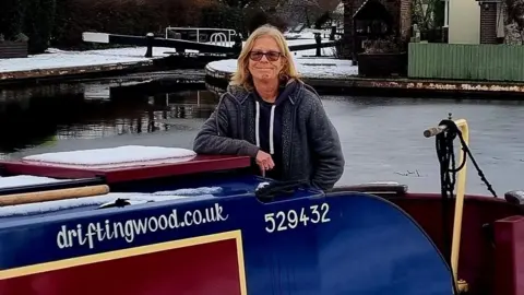 Laura Wood A blond woman in a grey hoodie is stood on the right of a blue and red canal boat with a canal lock in the background. The company name is written in white on the boat and there is a snow on the top of the boat. 