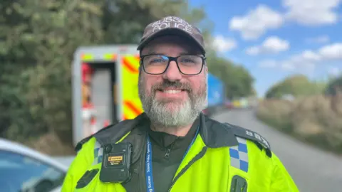 George Carden / BBC A man, Tom Carter, wearing a police uniform stood in a layby. He is standingin front of a van with hi-vis markings on it. He is smiling.