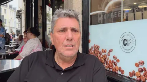 A person wearing a black polo shirt sits at an outdoor café table beside a large window displaying coffee bean graphics and the text “Coffee Republic” and “Roasted in Milan.” Several takeaway coffee cups are visible on nearby tables, with other café patrons seated in the background along a pedestrian street lined with buildings. The scene is photographed in daylight, with the seated person in sharp focus and the café surroundings clearly visible.