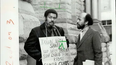 Santa signs prompt calls to police in Canadian city Toronto Star via Getty Images A black and white archival image of Richard Dildy. He is a black man with short black hair and a beard. He is wearing a formal button up shirt and a tie, with a coat on top. Around his neck hangs a sign that reads: "Down with Santa! Up with Truth! Stop Lying to the Kids!". He is looking right at the camera. To his left is another man looking at him.