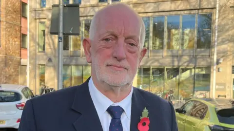BBC Deputy Steve Luce stood outside a large building with glasses panel windows. He is wearing a navy suit with a white shirt and a navy tie. He is looking directly at the camera and has a poppy on his jacket. There are a number of cars and bikes in the background.