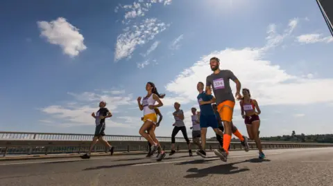 Blue sky and sunshine with light cloud above a groub of runners dressed in active wear and wearing race bibs 
