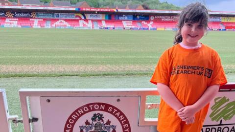 Heidi standing at Accrington Stanley football ground wearing an orange t-shirt with the Alzehimer's Research UK logo