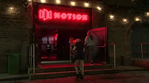 The entrance of Motion, with the word in red lights and a logo next to it. Women are walking in through the doors and it is night time.