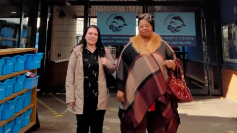 Women's Work Lab Reneta Atanasova standing next to a friend in front of the Dolphin School. She is wearing a beige coat over a black shirt.