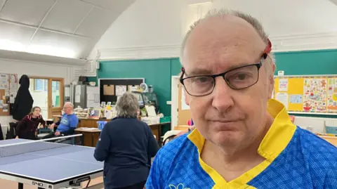 A man wearing a bright blue sports jersey with yellow chevron stripes and a logo that reads “Community Club Table Tennis” stands indoors near a table tennis set up. The room has green walls, a high curved ceiling, and several people in the background engaged in conversation and activities.