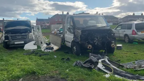 Three battered vehicles parked on a grass area with parts scattered across the field.