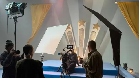 Eilís Doherty A behind the scenes shot showing people working at a blue stage. Gold curtains can be seen either side of the stage and there are gold stars behind the stage.