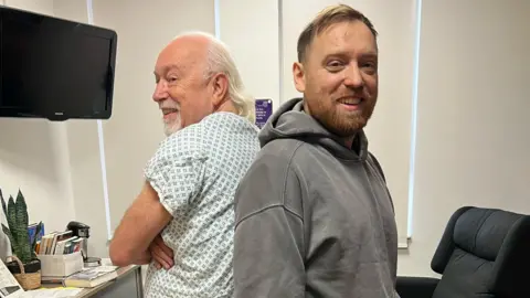 Supplied Two men stand back to back, looking at the camera. One, with white hair and a beard, in his 60s, is wearing a hospital patient gown. The other, in his 30s and with brown hair and a beard, is wearing a grey hoodie. They are in a room decorated white, with books and a plant on a surface against one wall, underneath a television. An armchair is on the other side of the room.