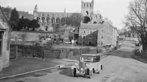 Marc Edwards A black-and-white image of an old car on an empty road with old buildings, including an abbey, in the background