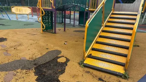 A yellow playground floor has a large chunk ripped out, revealing the earth beneath. There are some yellow steps leading to a slide. In the background you can see a desolate and run down play area. 