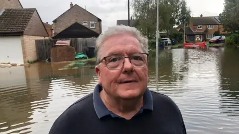 John Devine/BBC Michael Brown has short brown hair and dark rimmed glasses with a blue shirt under a dark coloured jumper. Behind him is a flooded housing estate.