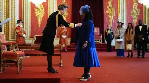 PA Media Dr Gee Walker stands in front of the Princess Royal who is placing the MBE on her blue blazer. Her family are watching in the background. The room is adorned with red and gold.