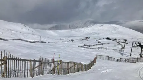 SAIS Glencoe Glencoe Mountain ski area is covered in snow. There is a snow fence and in the distance ski lift infrastructure. The sky above is grey and cloudy.