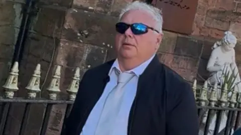 Allan Thursby arriving at court. He has short white hair and is wearing a black coat, a white shirt and grey tie. He is wearing black and blue sports-style sunglasses.