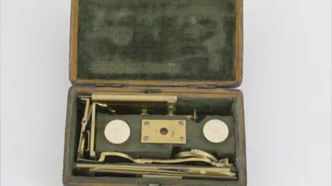 Kinghams Auctioneers A green velvet lined box, worn with age, open showing the brass instrument dismantled inside. Everything is neatly tucked into different sections of the box. 
