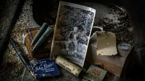 Angus Mackay Photography There is a collection of objects on a dusty floor. They include an old photograph of a man sitting on a wall, matchbooks, gun cartridges, a fountain pen and rusty bullets.