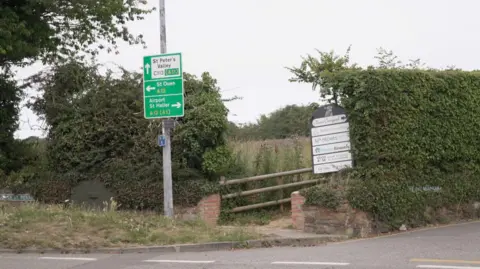 A rural road intersection with multiple directional signs. One green sign on a pole points to 'St Peter's Valley', 'St Ouen' and 'Airport, St Helier.' The area is surrounded by greenery, including a large hedge and overgrown vegetation. There is also a wooden gate next to the hedge leading into a field.