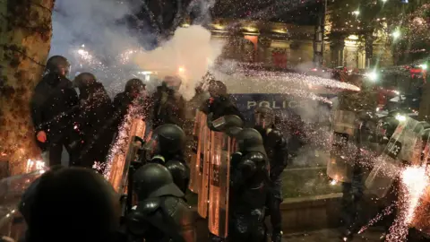 Reuters A firework explodes in a shower of yellow and red sparks above the heads of a group of riot police officers. They are wearing black uniforms and heavy riot helmets, their faces obscured. Several are holding clear plastic shields.