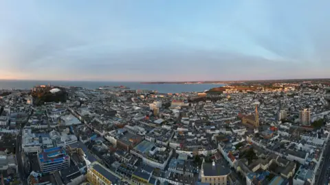 A fish eye lens view of St Hellier in Jersey showing churches housing and buildings and the sea in the background.
