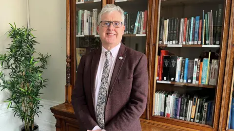 Philip Gerrard is a white-haired man with glasses in a red blazer and patterned tie with pink shirt. He is standing in front of a traditional glass and wood bookcase in an office.
