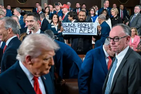 EPA/Shutterstock Al Grenne holds a sign that says "Black People Aren't Apes!" as Trump walks past in the chamber
