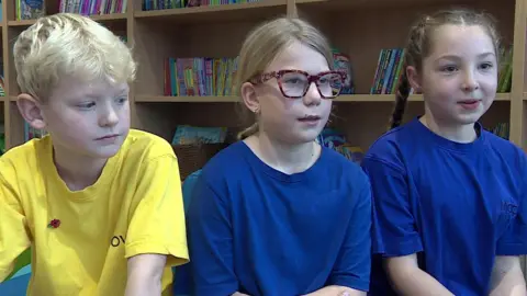 Image of three school children, two girls and a boy. The girls are wearing blue T-shirts and the boy is wearing a yellow top.  One of the girls has long brown hair in two plaits and the other girl has her hair tied back and is wearing a pair of glasses. The boy has short blonde hair. In the background can be seen shelves of books.