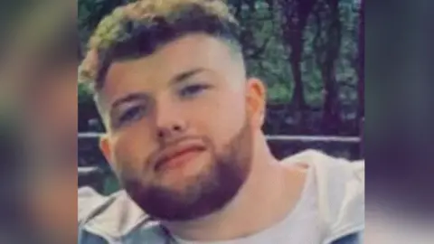 A young man with dark curly hair and a beard looks directly at the camera