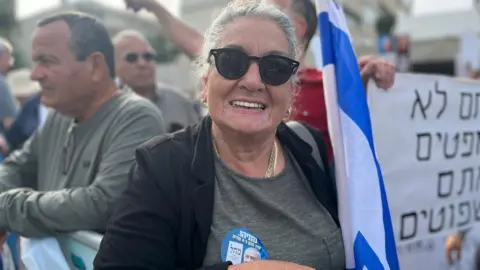 Eliza Ziv, een vrouw met grijs haar en een ronde zwarte zonnebril, glimlacht naar de camera. Ze houdt een opgevouwen Israëlische vlag vast en draagt ​​een sticker met het gezicht van Netanyahu erop.