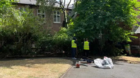 Offenders on the Immediate Justice scheme at Barker Gate Rest Garden in the city centre. Two people wearing high vis are facing shrubbery in the park with their backs to the camera. Gardening tools can be seen on the ground behind them. 