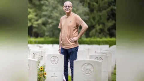 Venray War Cemetery Adoption Graves Foundation Theo Vervoort standing in the cemetery in Venray. He is wearing a light orange t-shirt and navy blue jeans. He is wearing glasses. He is smiling and has his hand on his hip.