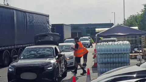 Michael Keohan/BBC A steady stream of cars queuing up to collect pallets of water at a carpark. There are two contractors, in yellow orange hi-vis jackets, standing beside the cars. 