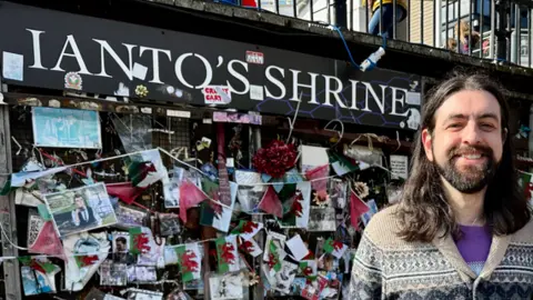 Golden Cat Studios A wall with the words Ianto's Shrine and hundreds of photographs and Welsh flags pinned to it. In front of it is a man with long black hair and a beard, wearing an Aztec print cardigan and smiling at the camera.