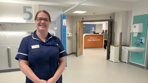 A woman in navy medical scrubs stands in a hospital ward, wearing glasses and a name badge. The setting is clean and clinical, with the number “5” marked on the wall behind and a sink unit to the side. Through an open doorway, a reception desk with a sign reading “Welcome to Oak Ward B” is visible.