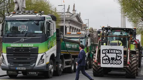 Vehicles parked on O'Connell Street in Dublin as protesters take part on the second day of a National Fuel Protest against rising fuel prices. In the foreground is a large haulage truck to the left of the picture and a tractor to the right. The tractor has a banner saying "No farms, no food". A man in a dark suit is crossing the road in front of the vehicles.