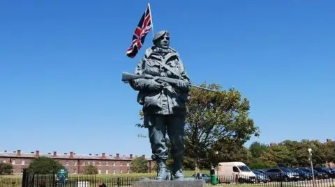A large bronze statue of a British soldier in the Falklands conflict is is shown with a union jack attached to the back against a clear blue sky. The figure holds a rifle and wears a large jacket.