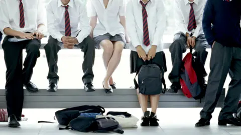 Getty Images A group of adolescents wearing school uniforms perch on a metal barrier. Only their legs and chests are in shot. They are wearing ties and holding rucksacks. 