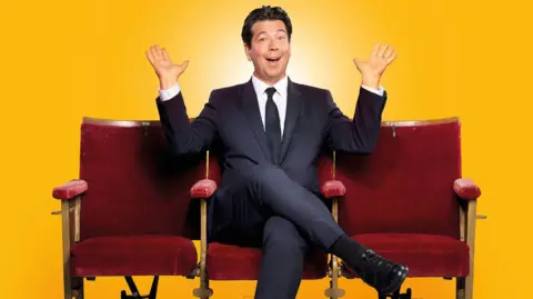 Michael McIntyre sitting cross-legged on a red chair with his hands in the air, posing for a publicity photo. The background is soft yellow. Two more red chairs are placed on either side of him. He is wearing a dark suit.