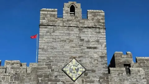 A close-up of the Castle Rushen Clock Tower, which is a large grey castellated structure with a Manx flag flying next to it.